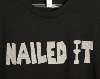 May include: A black short-sleeved t-shirt featuring the text NAILED IT printed in a silver glitter finish across the chest. The shirt has a crew neckline and a standard fit, with the silver lettering standing out against the solid black fabric.