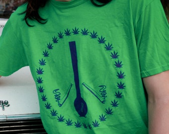 May include: A bright green short-sleeved t-shirt featuring a dark blue circular graphic. The design consists of a ring of small leaf icons surrounding a central vertical pipe illustration flanked by two smoking items, all printed on the chest area.