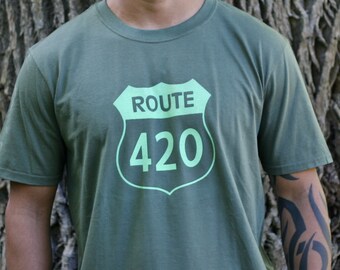 May include: A dark olive green short-sleeved t-shirt featuring a light green graphic of a highway shield shape with the text ROUTE 420 printed in the center. The shirt is paired with a charcoal gray knit beanie featuring a small diamond logo patch.