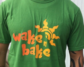 May include: A bright green short-sleeved t-shirt featuring a graphic print in orange and yellow. The design includes a stylized sun icon positioned above the text "wake n bake" written in a rounded, playful font across the center of the chest.