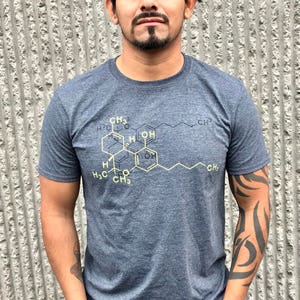 May include: A heathered dark blue short-sleeve t-shirt featuring a yellow chemical structure diagram printed on the chest. The graphic displays a complex molecular formula with various carbon and hydrogen labels, including CH3 and OH groups.