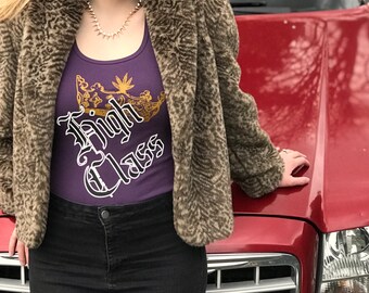 May include: A purple tank top featuring a gold crown graphic with a leaf detail and the white text High Class, worn under a brown leopard print faux fur jacket with black jeans and a gold spiked necklace, standing against a red vehicle.