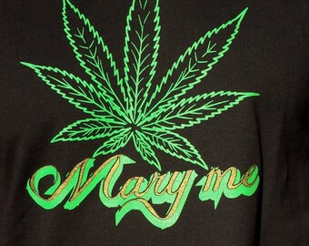 May include: A black t-shirt featuring a large, bright green graphic of a cannabis leaf centered above the text "Mary me" written in a stylized, cursive font with a gold-toned glittery texture and a bright green shadow effect.