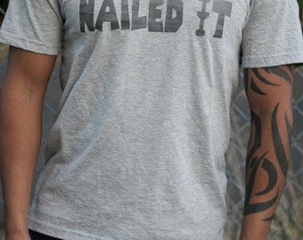 May include: A heather gray V-neck t-shirt featuring the text NAILED IT printed in a dark gray, blocky, stencil-style font across the chest. The shirt has short sleeves and a standard fit, constructed from a soft, lightweight fabric material.