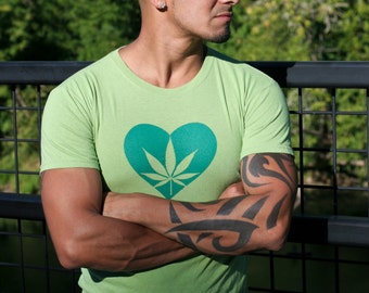 May include: A light green short-sleeved t-shirt featuring a dark green heart graphic on the chest. The heart contains a stylized cannabis leaf design centered within it. The shirt has a crew neckline and is made of a solid, lightweight fabric.