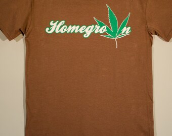 May include: A brown short-sleeve t-shirt featuring the white text Homegrown printed across the chest. The letter w is replaced by a green cannabis leaf graphic with a white outline. The shirt has a crew neckline and a standard fit design.