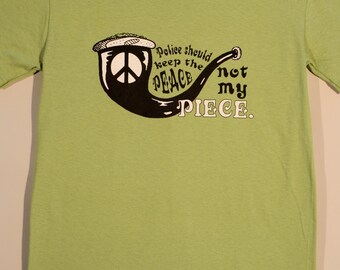 May include: A light green short-sleeve t-shirt featuring a black graphic of a smoking pipe with a peace sign inside the bowl. The text reads: Police should keep the PEACE not my PIECE. The design is centered on the chest of the crew neck shirt.