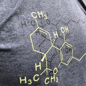 May include: A charcoal grey heathered t-shirt featuring a large, yellow chemical structure diagram printed on the front. The graphic displays complex molecular bonds with labels including CH3, OH, and H, layered over a faint black outline.