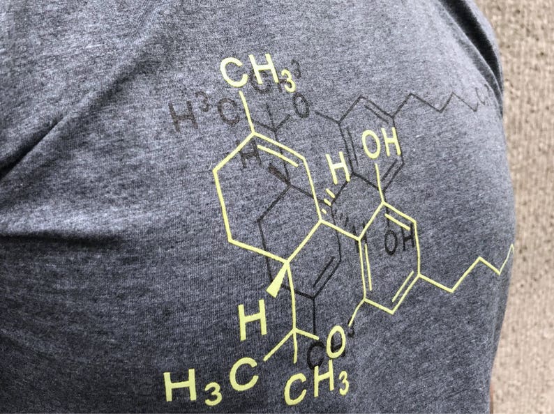 May include: A charcoal grey heathered t-shirt featuring a large, yellow chemical structure diagram printed on the front. The graphic displays complex molecular bonds with labels including CH3, OH, and H, layered over a faint black outline.