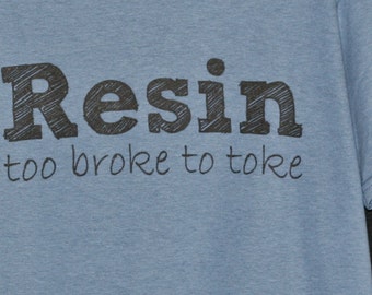 May include: A light blue t-shirt featuring the word Resin in a large, dark, sketch-style font, with the phrase too broke to toke printed underneath in a smaller, thinner, handwritten-style black font across the chest of the garment.