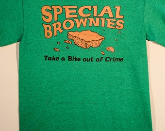 May include: A heather green short-sleeve t-shirt featuring the orange text SPECIAL BROWNIES above an illustration of a brownie piece with crumbs. Below the graphic, the black text reads Take a Bite out of Crime. The shirt has a crew neckline.