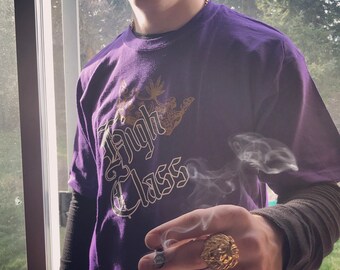 May include: A purple short-sleeved t-shirt featuring a gold graphic print of a crown and the text High Class. The shirt is worn over a black long-sleeved base layer. A gold-toned ring shaped like a lion head is worn on the finger.