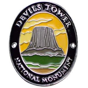 May include: A black and silver Devils Tower National Monument enamel pin with a yellow sunset background and a green base.