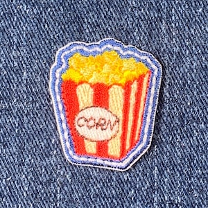 May include: A colorful popcorn patch with a red and white striped container, yellow popcorn, and the word "CORN" on a white circle. The patch has a blue border and is set against a blue denim background.
