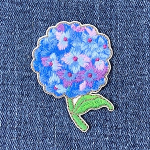 May include: Embroidered hydrangea patch featuring a cluster of blue, light blue, and purple flowers. The patch has a green stem and leaves, and is set against a blue denim background.