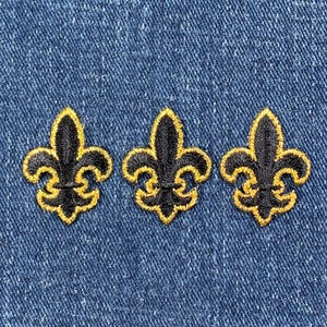 May include: Three black embroidered fleur-de-lis on a blue denim background.