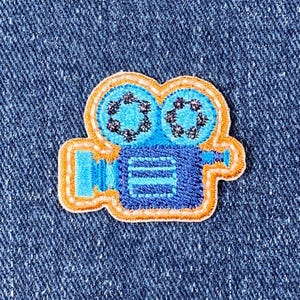 Peut inclure: Un patch brodé thermocollant représentant une caméra de cinéma vintage, en bleu et orange.