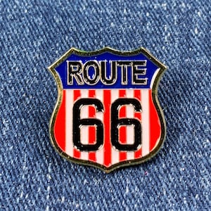 Könnte beinhalten: Ein rot-weiß-blauer Route 66-Pin mit goldfarbenem Rand. Der Pin ist schildförmig und trägt den Text "Route 66" in schwarzen Buchstaben.