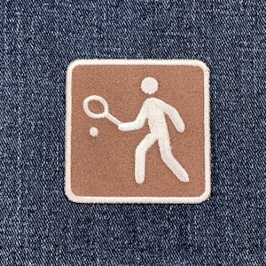 May include: A square, brown patch with a white border featuring a white silhouette of a person playing tennis. The person is holding a racket and hitting a ball. The patch is set against a blue denim background.