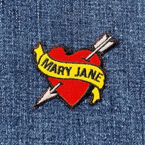 Peut inclure: Écusson brodé représentant un cœur rouge percé d'une flèche. Une bannière jaune sur le cœur porte l'inscription "MARY JANE" en lettres blanches. L'écusson est posé sur un fond en denim bleu.