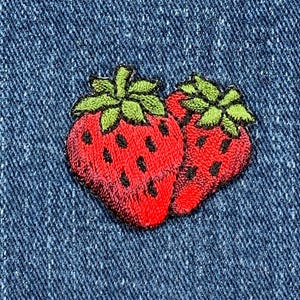 May include: Two embroidered strawberries on a blue denim background. The strawberries are red with green leaves and are stitched in detail.