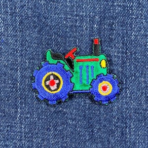 May include: Embroidered patch of a green tractor with blue wheels and a red steering wheel. The tractor has a yellow headlight and a black smokestack. The patch is on a blue denim background.