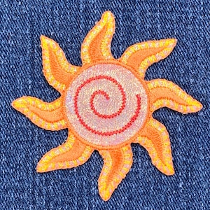 May include: A bright orange sun with a pink and red spiral in the centre. The sun is embroidered with a sparkly finish and has a denim background.