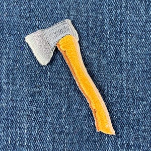 May include: Embroidered patch of a small axe with a grey head and an orange handle. The patch is on a blue denim background. The axe is a simple, stylized design.