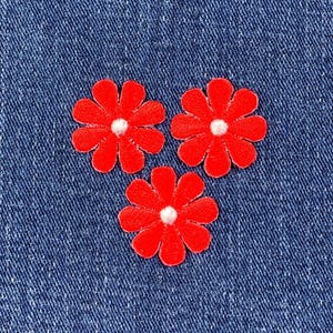 Könnte beinhalten: Drei rote Gänseblümchen-Blumenaufnäher mit weißen Zentren sind auf einem blauen Denim-Hintergrund angeordnet. Die Blumen haben abgerundete Blütenblätter und sind gleichmäßig verteilt. Der Denim hat ein strukturiertes Aussehen.