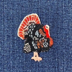 May include: A colorful turkey patch with a detailed design. The patch features a black body with white, orange, and blue accents, a red wattle, and a vibrant red and orange tail. The patch is set against a blue denim background.