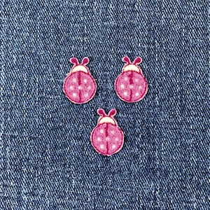 May include: Three pink ladybug-shaped embellishments with white polka dots and a white head, set against a blue denim background. These decorative elements are ideal for crafting or adding a whimsical touch to clothing.