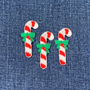 May include: Three embroidered candy cane patches. Each candy cane has red and white stripes, a white curved top, and a green bow. The patches are on a blue denim background.