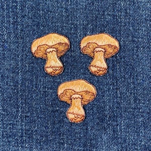 以下が含まれることがあります： 青いデニムの背景に、刺繍された茶色のキノコのパッチが3つある。