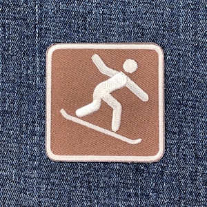 May include: A square patch featuring a skier in white on a brown background. The patch has a white border and is set against a blue denim fabric.