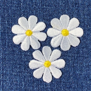 Peut inclure: Trois patchs de fleurs de marguerite blanches avec des centres jaunes sur un fond de denim bleu.
