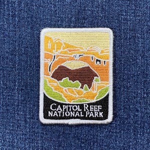 May include: Embroidered patch featuring a landscape scene from Capitol Reef National Park. The patch has a rectangular shape with a white border. The design includes a brown rock formation, green vegetation, and the text "CAPITOL REEF NATIONAL PARK".