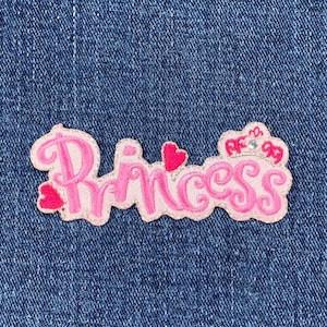 May include: A pink embroidered patch with the word "Princess" in a cursive font. The patch features a crown and hearts, and is set against a blue denim background.