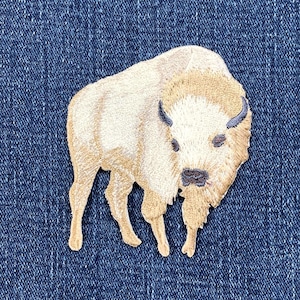 May include: Embroidered patch of a beige bison with a tan head and dark blue horns, set against a dark blue denim background. Suitable for clothing or accessories, this patch is likely for decorative purposes.