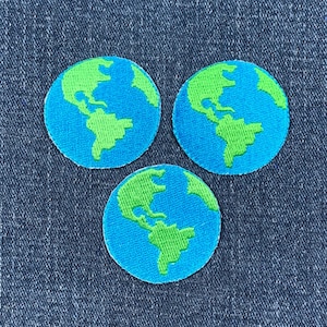 May include: Three circular embroidered patches featuring a blue globe with green continents. The patches are arranged on a blue denim background. The patches are likely for clothing or accessories.