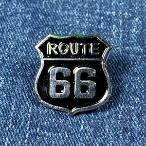 May include: A silver metal pin shaped like a Route 66 highway sign. The sign is black with white lettering and the number 66.