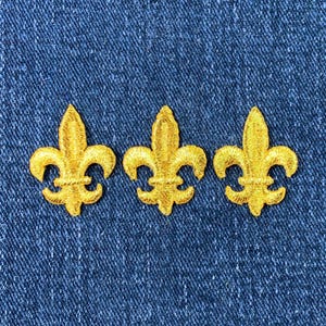 Peut inclure: Trois écussons dorés en forme de fleur de lys sur un fond en denim bleu. Chaque écusson présente un motif brodé détaillé de la fleur de lys stylisée, symbole de la royauté française. Les écussons sont espacés uniformément, mettant en valeur leurs détails complexes.