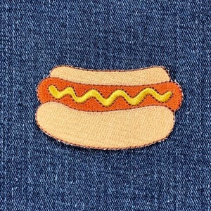 May include: Embroidered patch of a hot dog with a yellow zigzag of mustard on a light orange hot dog and bun. The patch is outlined in a darker orange thread. The background is a textured blue fabric.