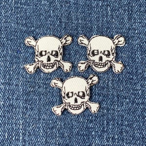 Könnte beinhalten: Drei weiße gestickte Totenkopf- und Knochenkreuz-Patches auf einem blauen Jeans-Hintergrund.