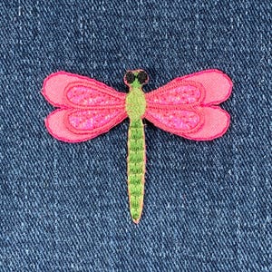 May include: A pink and green dragonfly patch on a blue denim background. The dragonfly has pink wings with a textured pattern, a green body, and black eyes. The patch is embroidered.