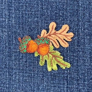 May include: Embroidered patch featuring two acorns and an oak leaf in shades of brown, orange, green, and yellow. The patch is on a blue denim background.