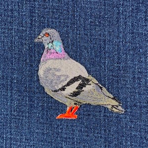 May include: A denim fabric with an embroidered pigeon design. The bird is gray with black wing accents, a pink and blue neck, and orange feet. The background is a textured, dark blue denim.