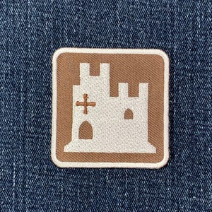 May include: A square patch featuring a brown castle design with a white outline and a white cross. The patch is set against a blue denim background.