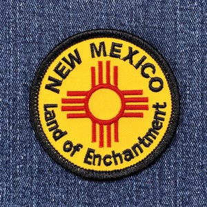 May include: A round patch featuring the New Mexico state flag design. The patch has a yellow background with a red Zia symbol in the center. The words "NEW MEXICO" and "Land of Enchantment" are in black text around the edge.