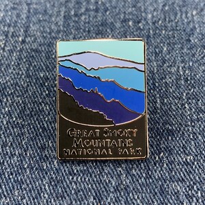 May include: Rectangular enamel pin featuring a landscape design of the Great Smoky Mountains National Park. The pin has a black border and the text "Great Smoky Mountains National Park". The landscape design includes shades of blue, purple, and teal.