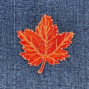 Könnte beinhalten: Gestickter orangefarbener Ahornblatt-Aufnäher auf blauem Jeansstoff. Das Blatt hat detaillierte Stiche mit einem helleren orangefarbenen Innenraum und einer dunkleren orangefarbenen Kontur. Der Aufnäher ist ein dekoratives Element für Kleidung oder Accessoires.